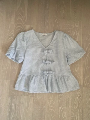 Blårandig blus med rosetter Gina Tricot - Supersöt blårandig blus från Gina Tricot med puffärmar, v-ringning och tre dekorativa rosetter framtill. Blusen har volang nedtill och är gjord i ett lätt bomullstyg som passar perfekt till sommaren. Perfekt för dig som gillar gulliga detaljer och en luftig stil.