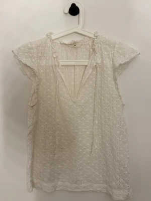 Vit broderad blus  - Söt vit blus med broderade hålmönster och volangkant vid halsen. Blusen har korta ärmar och knytband fram i urringningen. Tillverkad i ett lätt och luftigt tyg, perfekt för varma dagar! 