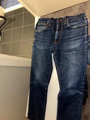 Blå jeans från Nudie Jeans - Säljer ett par klassiska blå jeans från Nudie Jeans med snygga slitningar och kontrastsömmar. Jeansen har fem fickor, normal midja och raka ben. Bakfickorna har Nudie Jeans ikoniska broderade vågmönster. Perfekta till en avslappnad stil. W 31 passar w29