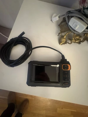 Inspektionskamera med display och lång kabel - Säljer en svart inspektionskamera med stor färgdisplay och lång flexibel kabel. Kameran har knappar för navigering och power, samt orange detaljer. Perfekt för att undersöka trånga utrymmen och svåråtkomliga platser.