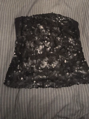 Svart paljett-tubtop - Säljer en svart tubtop täckt med glittrande paljetter i storlek 38.  Det följer med axelband, den är i helt nytt skick med prislappen kvar. Den är streshig.