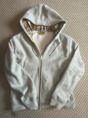 Grå zip hoodie från Burberry - Snygg grå hoodie med dragkedja från Burberry. Klassisk huva med det ikoniska rutmönstret på insidan och broderad logga på bröstet. Tillverkad i mjuk bomullsblandning, ribbade muddar och fickor framtill. Perfekt för en chill och stilren look.