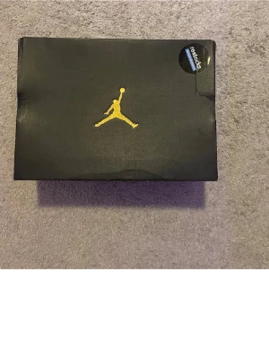 Air Jordan 1 High svart/guld/vit - Säljer ett par Air Jordan 1 High sneakers i svart, guld och vitt. Klassisk high-top siluett med svart och gulddetaljer, vit tåbox och sula. Skorna har den ikoniska Jordan-loggan och är tillverkade i läder för en clean och sportig look.