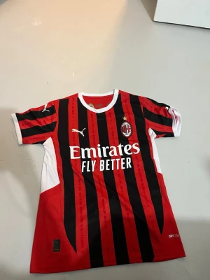 AC Milan matchtröja Puma röd/svart - Säljer en officiell AC Milan matchtröja från Puma med klassiska röda och svarta vertikala ränder, vita detaljer och klubbmärke på bröstet. Tröjan har korta ärmar, rund hals och tryck med 'Emirates Fly Better' på framsidan. Perfekt för fotbollsfans!