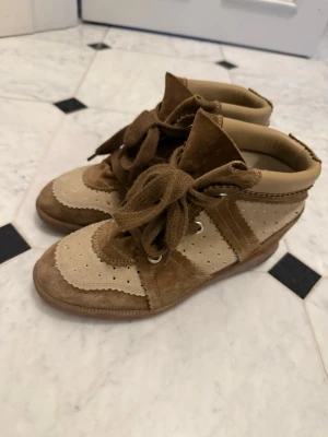 Beige sneakers Isabel Marant - Snygga beige sneakers från Isabel Marant med högt skaft, perforerade detaljer och breda bruna skosnören. Skorna är gjorda i mocka och textil, med gummisula och coola kontraster mellan ljusa och mörka partier. Perfekta för en trendig och avslappnad look.