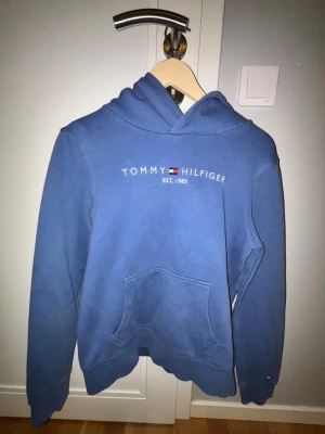 Blå hoodie från Tommy Hilfiger - Snygg blå hoodie från Tommy Hilfiger med klassisk logga och texten 'EST. 1985' på bröstet. Hoodien har en stor känguruficka framtill och en skön huva. Perfekt för dig som gillar streetwear och vill ha en stilren look.