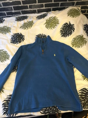 Blå halvzip från Polo Ralph Lauren - Snygg blå halvzip-tröja från Polo Ralph Lauren med klassisk broderad logga på bröstet. Har ej använt den mycket max 5 gånger. Inget slitage på den alls och väldigt skön.