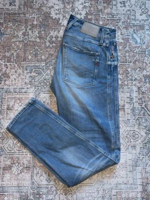 Blå raka jeans från Replay - Säljer ett par klassiska blå jeans från Replay med raka ben och femficksmodell. Jeansen har snygga slitningar och en ljus tvätt framtill för en avslappnad look. Tillverkade i bomull med den ikoniska Levi's-lappen bak i midjan.