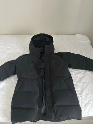 Svart vinterjacka från Sail Racing - Säljer en svart vinterjacka från Sail Racing med huva och dragkedja framtill. Jackan har två bröstfickor med dragkedja och två sidofickor. Perfekt för kalla vinterdagar och har en stilren, enkel design.