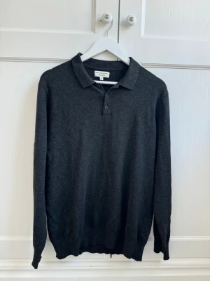 Mörkgrå pikétröja  - Mörkgrå långärmad pikétröja från Christiano Baldinucci. Tröjan har klassisk krage, tre knappar vid halsen och ribbade muddar vid ärmslut och nederkant. Tillverkad i mjukt material som känns skönt mot huden.