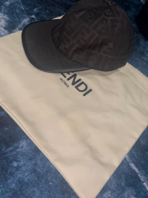 Svart keps från Fendi med FF-mönster - Snygg svart keps från Fendi med det klassiska FF-monogrammet över hela kepsen. Kepsen har böjd skärm med svart kant och är tillverkad i ett tygmaterial som känns lyxigt. Perfekt för dig som vill ha en exklusiv accessoar med streetkänsla.