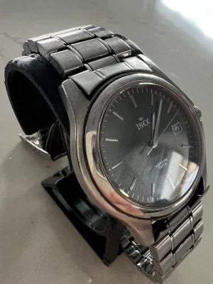 Klassisk silverfärgad INEX klocka - Stilren armbandsklocka från INEX med rund svart urtavla, datumvisning och markerade index. Boetten och länken är i silverfärgad metall vilket ger en tidlös look. Saphirglas för extra tålighet. Perfekt accessoar för dig som gillar klassisk stil.