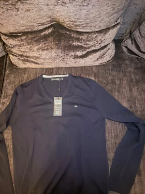 Mörkblå stickad tröja J.Lindeberg - Snygg mörkblå stickad tröja från J.Lindeberg med V-ringning och diskret logga på bröstet. Tröjan har långa ärmar och är tillverkad i mjukt material som passar perfekt till lager på lager. Enkel och stilren design som funkar till det mesta.