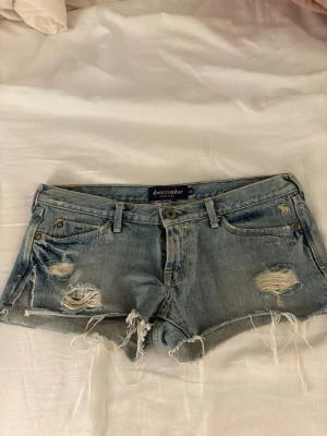 Ljusa jeansshorts från Abercrombie - Säljer ett par ljusblå jeansshorts från Abercrombie med slitna detaljer och fransiga kanter. Klassisk femficksmodell med låg midja. Perfekta för en avslappnad och trendig sommarlook.