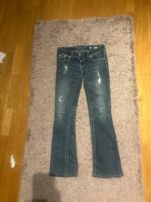 Miss Me bootcut jeans med brodyr - Säljer ett par blå bootcut jeans från Miss Me med slitningar och snygga broderade detaljer på bakfickorna. Jeansen har låg midja, kontrastsömmar och klassisk femficksdesign. Perfekta för dig som gillar en avslappnad men ändå trendig look. Dragkedjan dock lite trasig. Pros kan såklart diskuteras. Skriv privat för fler bilder