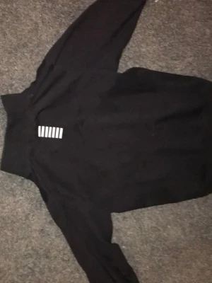 Svart EA7 hoodie med dragkedja - Snygg svart hoodie från EA7 Emporio Armani med dragkedja framtill och vit EA7-logga på bröstet. Har vita ränder som detalj på fickan och hög krage. Perfekt för en sportig och avslappnad stil. Tillverkad i mjukt material för extra komfort.