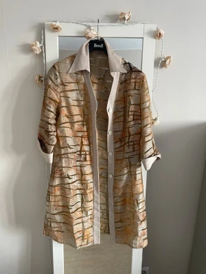 Beige mönstrad  lång jacka  med knappar - Unik beige lång jacka -skjorta  med abstrakt mönster i bruna och orange toner. Skjortan har knappar framtill, klassisk krage och trekvartsärm med breda manschetter. Perfekt för dig som vill sticka ut med en cool och konstnärlig vibe.
