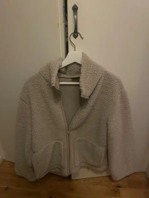 Beige teddyjacka med dragkedja - Mysig beige teddyjacka med hög krage och dragkedja framtill. Jackan har två stora fickor och är gjord i fluffigt, mjukt material som ger en avslappnad och trendig look. Perfekt för kyliga dagar när du vill vara både varm och snygg.