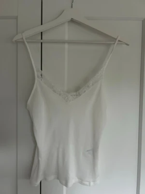 Vit linnetopp med spetsdetalj - Säljer en vit linnetopp med tunna axelband och v-ringning dekorerad med spets. Toppen har en enkel och stilren design som passar perfekt till sommaren. Materialet känns mjukt och lätt mot huden.
