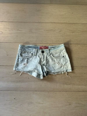 Ljusblå jeansshorts  - Säljer ett par ljusblå jeansshorts med slitna detaljer och fransig kant. Shortsen har fem fickor, knappgylf och klassisk passform. Perfekta för varma dagar och en avslappnad stil. Storlek 26