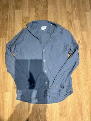 Blå linneskjorta från Knowledge Cotton - Säljer en stilren blå skjorta från Knowledge Cotton Apparel i mjukt linne. Skjortan har klassisk krage, långa ärmar och vita knappar framtill. Perfekt för dig som gillar en avslappnad men snygg look.