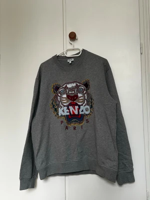 Grå sweatshirt från Kenzo - Säljer en grå sweatshirt från Kenzo. Tröjan har rund halsringning, långa ärmar och ribbade muddar. Perfekt statement-plagg för dig som gillar streetwear och snygga detaljer.