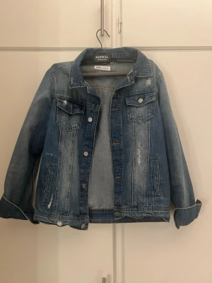 Blå jeansjacka - Klassisk blå jeansjacka med slitna detaljer och två bröstfickor med lock. Jackan har knäppning framtill, raka ärmar och en avslappnad passform. Den är i Storlek 164, men jag själv har använt den trots att jag är äldre, är i princip i xs/s.