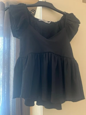 Svart volangblus med v-ringning - Supersöt svart blus med v-ringning och volangdetaljer. Toppen har korta ärmar med volang och en luftig, flowy passform. Perfekt för dig som gillar en feminin och trendig stil.