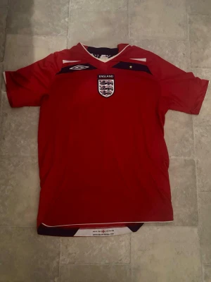 Äkta England Beckham matchtröja Medium - Säljer en röd England fotbollströja från Umbro med Beckham och nummer 7 på ryggen. Tröjan är inte använd en endaste gång. Storlek Medium. Ikonisk fotbollsspelare.