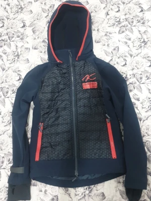 Mörkblå vindjacka från Equestrian Life - Sportig mörkblå vindjacka med huva från Equestrian Life. Jackan har röda detaljer, hel dragkedja framtill och två röda dragkedjefickor. Framsidan har ett diskret hexagonmönster och logga på bröstet. Perfekt för dig som gillar en aktiv stil.