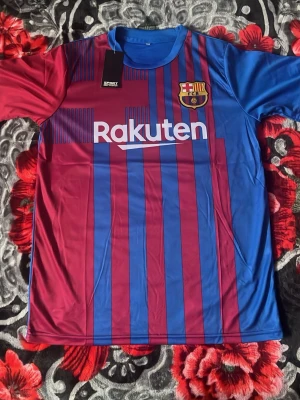 Barcelona matchtröja blå/röd 21-22 - Snygg Barcelona matchtröja i blått och rött med klubbmärke och Rakuten-tryck på bröstet. Tillverkad i lätt och ventilerande polyester, perfekt för fotboll eller träning. Klassiska vertikala ränder och rund hals. Kortärmad modell.
