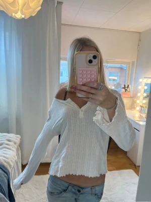 Vit ribbad offshoulder topp - Säljer en vit ribbad topp med offshoulder-design och långa ärmar. Toppen har knappar framtill och vågig kant nertill och vid ärmsluten. Perfekt för dig som gillar en avslappnad men ändå trendig stil.