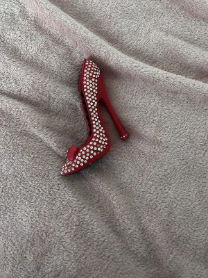 Röd smyckeshållare i form av klack - Snygg smyckeshållare formad som en röd högklackad sko med glittrande strass och en liten rosett framtill. Perfekt för att förvara ringar eller små örhängen. Materialet är hårdplast med mjuk svart insida för smyckena.
