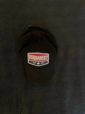 Svart keps från Dsquared2 - Säljer en svart keps från Dsquared2 med broderad logga i rött och vitt framtill. Kepsen har böjd skärm och justerbar rem bak. Tillverkad i bomullsmaterial och har mesh på insidan för extra komfort. Perfekt för dig som vill ha en stilren och trendig accessoar.