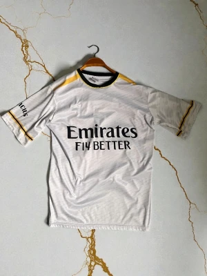 Real Madrid matchtröja Vinicius Jr - Snygg vit Real Madrid matchtröja med svarta och gula detaljer. Tryck med 'Emirates Fly Better' på framsidan och 'Vinicius 20' på ryggen. Korta ärmar och sportig passform, perfekt för fotboll eller träning.