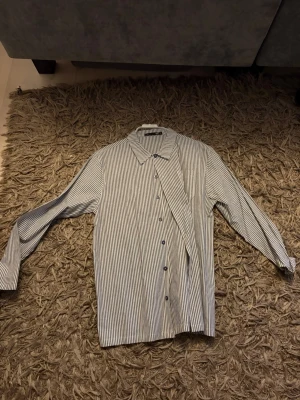 Randig skjorta med knappar - Klassisk vit skjorta med tunna svarta ränder och knappar framtill. Skjortan har lång ärm och en rak passform, perfekt för en clean och stilren look. Tillverkad i mjukt bomullstyg som känns skönt mot huden. (Aldrig använd)