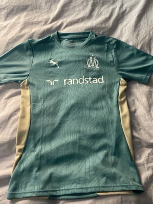 Turkos Marseille matchtröja Puma - (Köpt i sportaffär när jag va i Marseille sommaren 2025) Snygg turkos matchtröja från Olympique de Marseille, tillverkad av Puma. Tröjan har vita och beige detaljer, klubbmärke och Puma-logga på bröstet samt sponsortryck. Tillverkad i lätt och ventilerande material, perfekt för träning eller match.