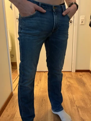 Blå raka jeans från Lee - Snygga blå jeans från Lee med klassisk rak passform och fem fickor. Jeansen har en djupblå tvätt och tydliga sömmar, samt Lee-logga på bakfickan och läderpatch i midjan. Perfekta för en avslappnad och stilren look.