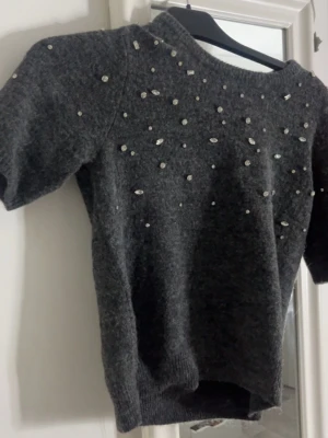 Mörkgrå stickad tröja med pärlor - Snygg mörkgrå stickad tröja med korta ärmar och glittrande pärlor och stenar på framsidan. Tröjan har rund halsringning😊aldrig använd