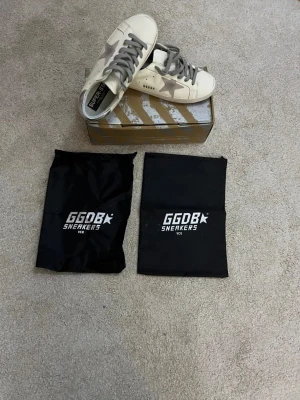 Golden Goose Superstar sneakers - Golden Goose Superstar sneakers i vit med grå stjärndetalj på sidan, silverfärgad häl och grå snörning. Skorna har en platt sula och rund tå. Materialet är skinn och mocka. Medföljer originalkartong och två svarta dustbags med GGDB-logga. Skorna är I storlek 41 och pris kan självklart diskuteras! 