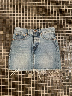 Blå jeanskjol med fransig kant - Snygg blå jeanskjol i klassisk modell med fem fickor och rå, fransig nederkant. Kjolen har knapp och dragkedja framtill och är perfekt för en avslappnad stil. Passar till det mesta och ger en cool vibe.