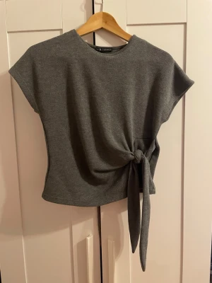 Zara knyt topp - Snygg grå stickad topp med korta ärmar och trendigt knyt i sidan. Toppen har en enkel, rak passform och är perfekt för dig som gillar stilrena plagg med en twist. Materialet känns mjukt och bekvämt mot huden.