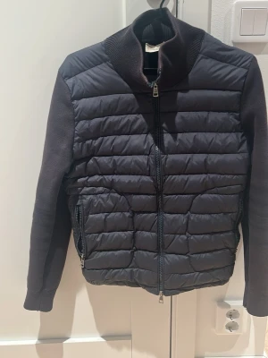 Mörkblå Moncler cardigan med stickade ärmar - Säljer en mörkblå cardigan från Moncler med quiltad front och stickade ärmar. Jackan har hög krage, dragkedja framtill och två fickor med dragkedja. Klassisk Moncler-logga på ärmen och en clean, modern look. Perfekt för dig som vill ha både värme och stil. Den är solblekt men går troligen att lösa genom en kemtvätt osv. Köpt här på Plick. Den har även 2 små hål vid ärmslutet men går och laga utan problem hemma eller hos en skräddare 