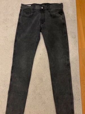Mörk gråa slim fit jeans från Replay - Säljer ett par svarta slim fit jeans från Replay med klassisk femficksdesign och snygga detaljer på bakfickorna. Jeansen har knappgylf och är tillverkade i stretchigt denimtyg för extra komfort. Perfekta för en trendig och avslappnad look.