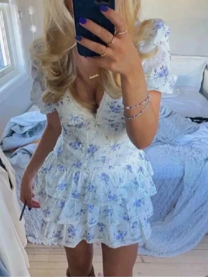 Trendig klänning - Supersöt vit klänning med blå blommönster och volanger. Puffärmar och v-ringad hals ger en romantisk vibe. Tillverkad i mjuk bomull, perfekt för varma dagar. Spetsdetaljer längs sömmarna och figurnära passform.