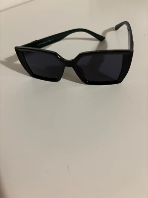 Solglasögon - Snygga svarta cat eye solglasögon med mörka glas och klassiska gröna och röda detaljer på skalmarna. Guldigt emblem på sidan ger en lyxig touch. Perfekta för dig som vill sticka ut med en trendig accessoar. Finns ingen kartong till.