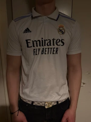 Real Madrid matchtröja Adidas vit - Snygg vit Real Madrid matchtröja från Adidas med klubbmärke och Adidas-logga på bröstet. Tröjan har korta ärmar, krage och trycket 'Emirates Fly Better' på framsidan. Perfekt för dig som älskar fotboll och vill representera ditt favoritlag.