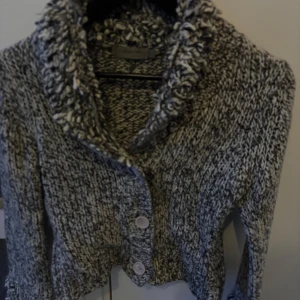 Grågrönmelerad stickad kofta med krage - Säljer en grågrön melerad stickad kofta med stora knappar framtill och en bred, fluffig krage. Koftan har lång ärm och är gjord i ett tjockt, mjukt garn som ger en mysig känsla. Perfekt för kyliga dagar och enkel att matcha till olika outfits.