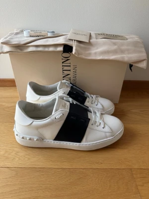 Valentino open skor  - Snygga vita sneakers från Valentino Garavani med bred svart läderdetalj över mitten och ikoniska nitar på hälen. Skorna har klassisk låg profil, vita skosnören och är tillverkade i premiumläder. Perfekta för dig som vill ha en clean men ändå lyxig look.