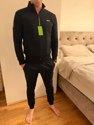 Svart tracksuit från BOSS - Snygg svart tracksuit från BOSS med dragkedja framtill och hög krage. Setet består av en jacka med diskret logga på bröstet och matchande byxor med dragsko i midjan. Perfekt för träning eller chill. Materialet är mjukt och stretchigt för maximal komfort.